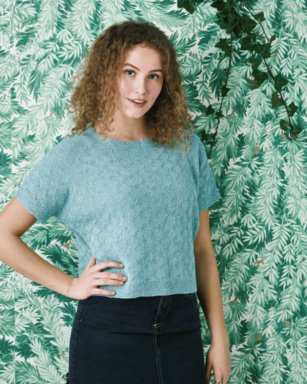 Kvinde med krøllet hår iført lyseblå strikbluse og mørkt nederdel, stående foran en grøn plantebaggrund.