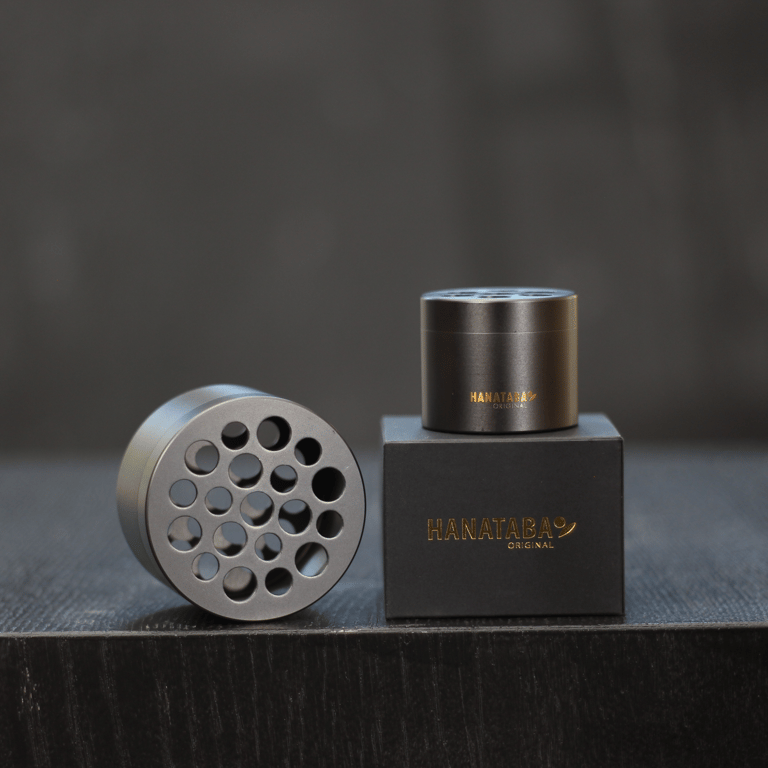 Sort metal urtekværn med huller, placeret ved siden af en sort æske med guldskrift "HANATABA ORIGINAL".