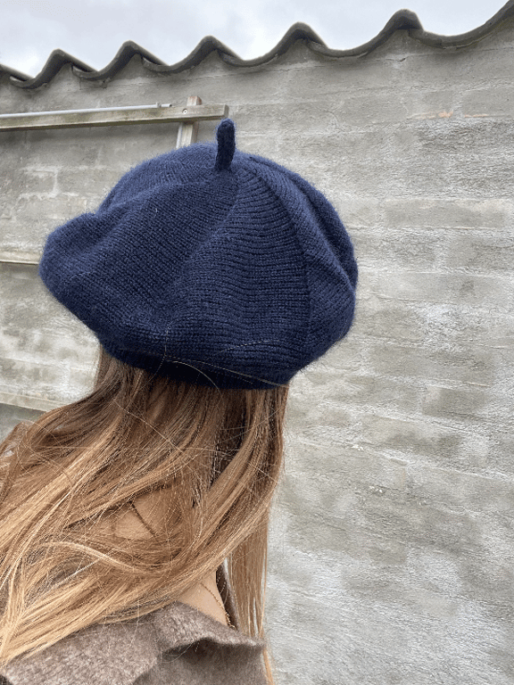 Kvinde set bagfra med langt lyst hår iført en mørkeblå strikket baret, foran en grå murstensvæg.