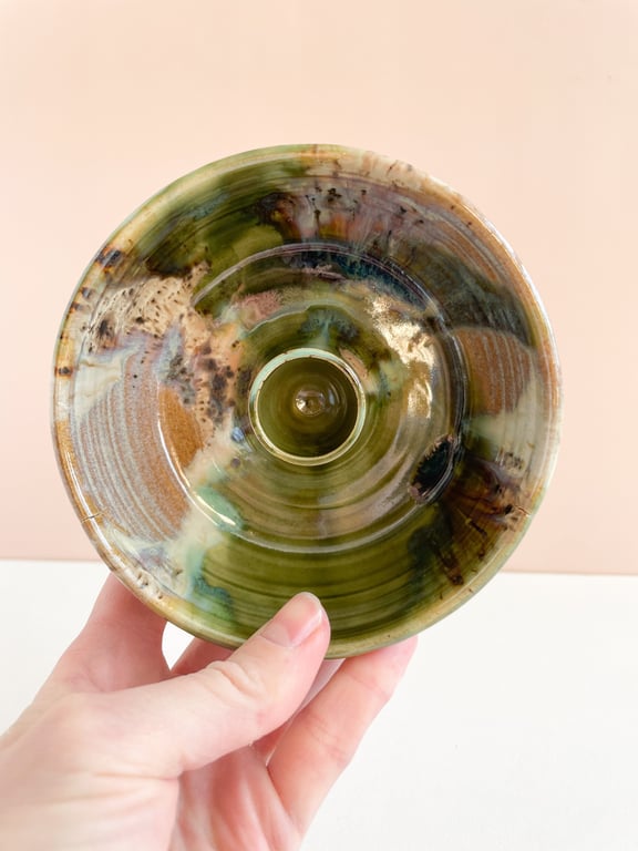 Hånd, der holder en glasurbelagt keramisk skål eller lysestage med grønne, brune og beige nuancer.