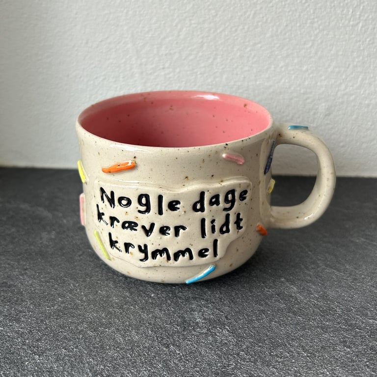 Kop med beige yderside, pink inderside og tekst på dansk "Nogle dage kræver lidt krymmel" med farverige krymmel-dekorationer på ydersiden.