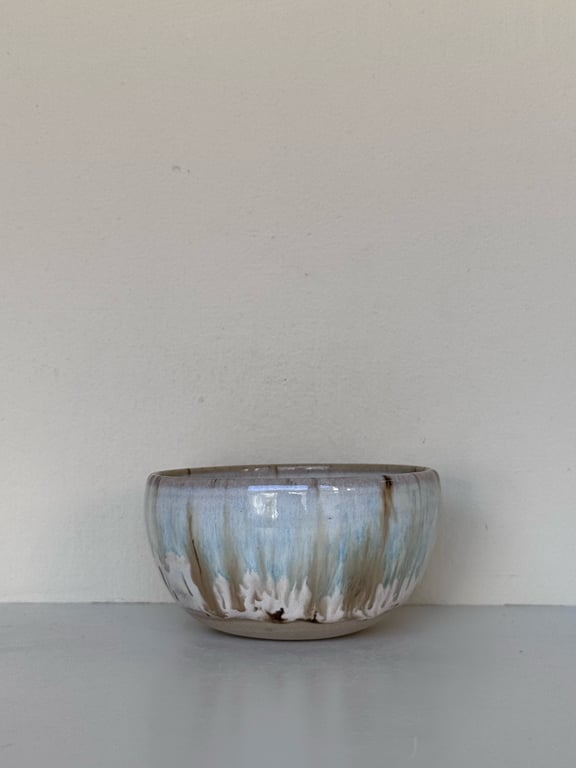 En keramisk skål med glasur i blå, beige og brune nuancer, placeret på en lys overflade foran en hvid væg.