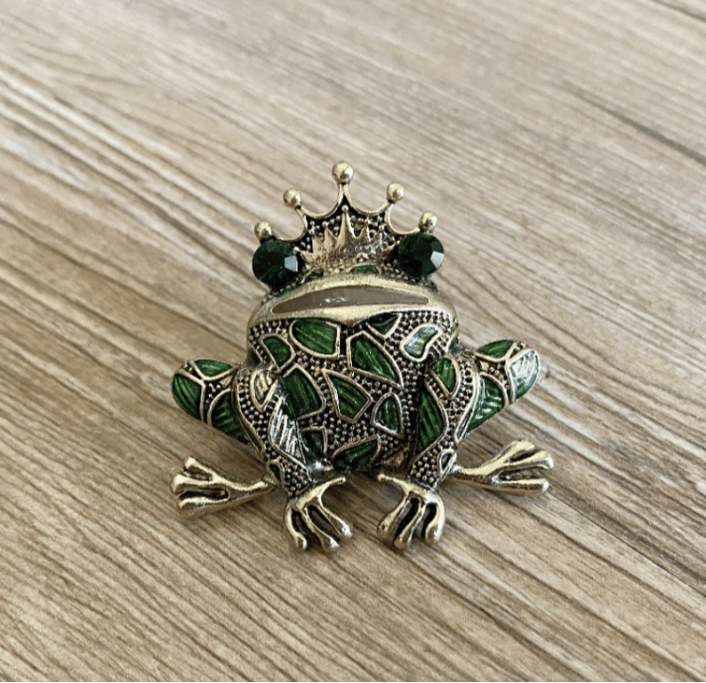 Brosje formet som en frø med grønne emaljedetaljer og en krone på hovedet, placeret på en lys træbordplade. (2)