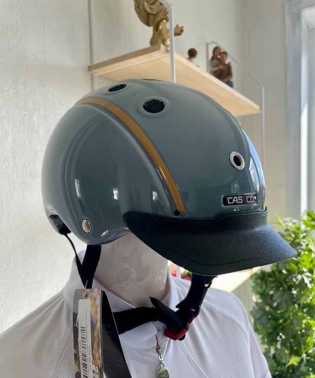 Grå hjelm med mørk skygge og sorte stropper, placeret på en hvid mannequin eller buste indendørs med hylder og grønne planter i baggrunden.