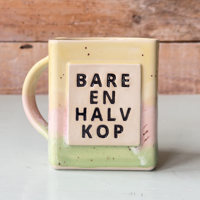 Keramikkop med firkantet form og teksten "Bare en halv kop" på dansk, i gule, lyserøde og grønne nuancer, placeret på et lyst bord med en træbaggrund.