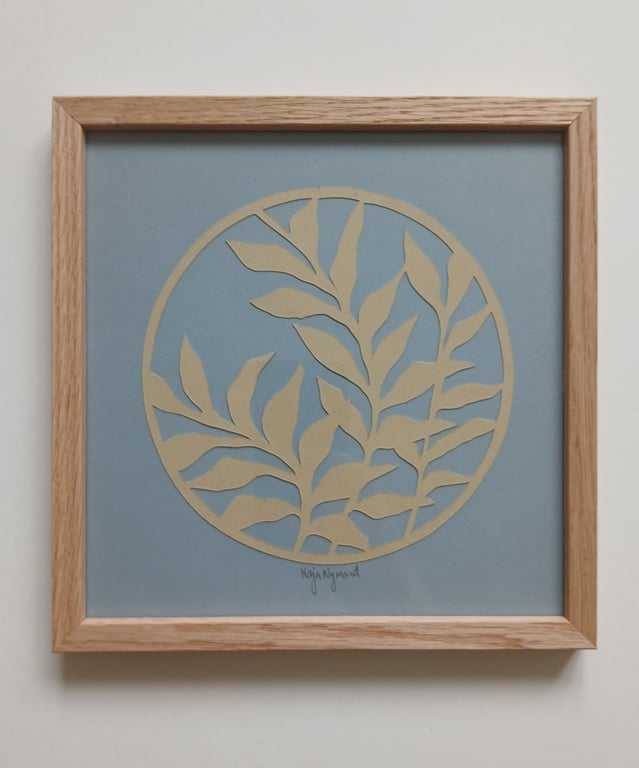 Indrammet papirkunst med beige bladformer arrangeret i en cirkel på en blå baggrund.