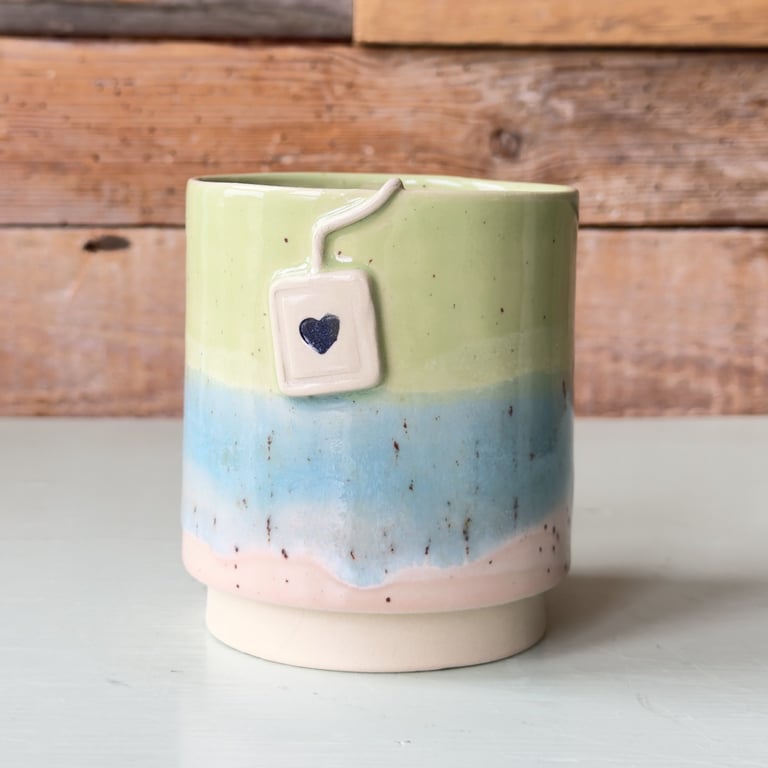 Keramisk kop med farverne grøn, blå og lyserød, dekoreret med en porcelæns-tepose med et hjerte.