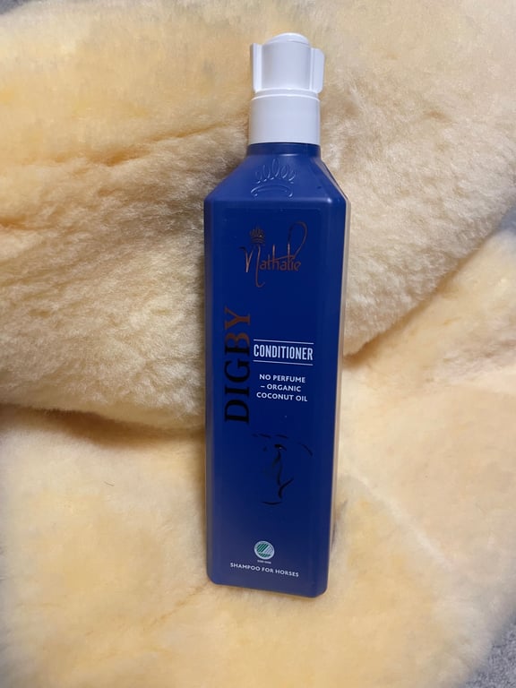 Blå flaske med hvid pumpe, mærket "Digby Conditioner" med kokosolie og uden parfume, placeret på en cremefarvet uld- eller pelsagtig baggrund.