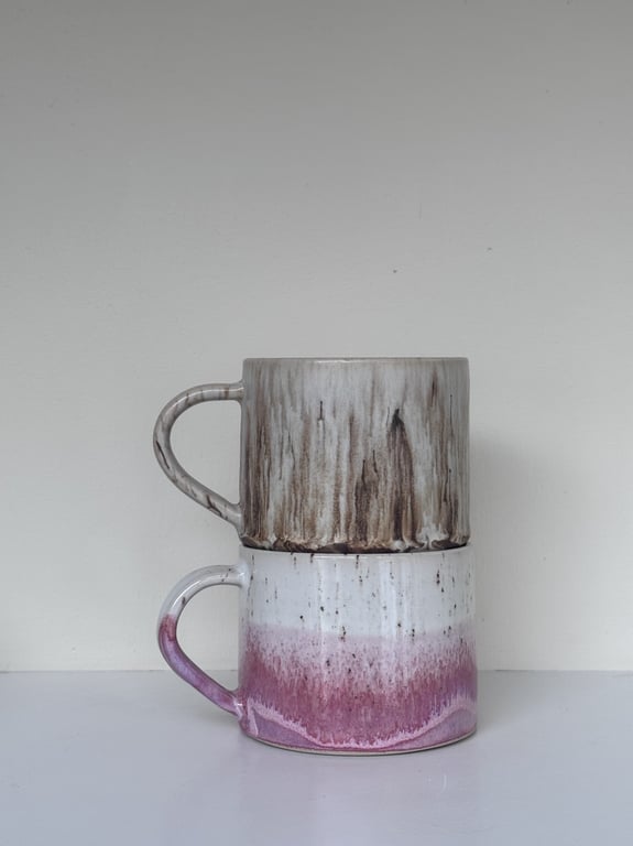 To keramiske krus med håndtag, det øverste har en gråbrun glasur, det nederste har en hvid og lyserød glasur, stablet på hinanden mod en hvid baggrund.