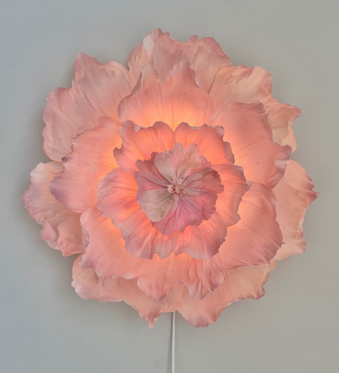 Stor lyserød blomsterlampe med bløde, bølgede kronblade, der lyser varmt indefra, monteret på en lys grå væg.