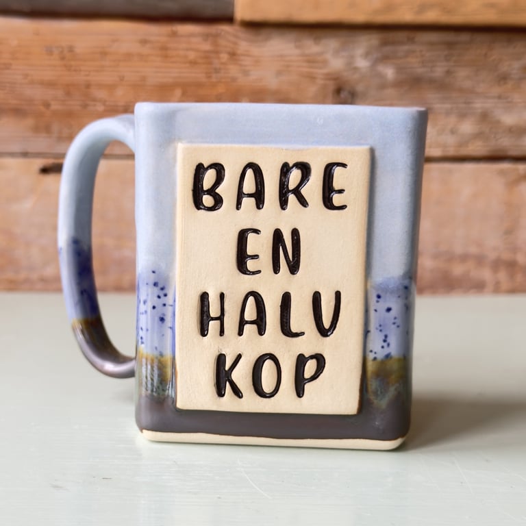 Grå og beige keramisk kop med teksten "BARE EN HALV KOP" i sort reliefbogstaver.