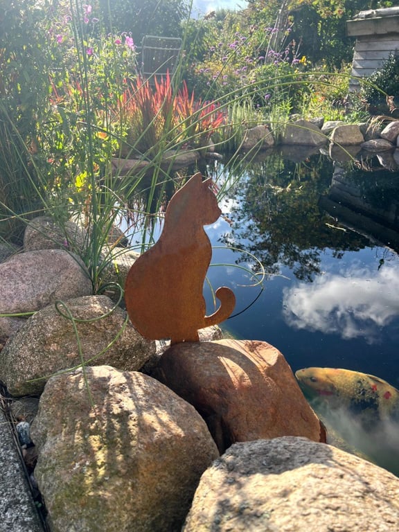 En metalstatue af en kat placeret på sten ved kanten af en dam med spejling af himlen og en koi-fisk i vandet.