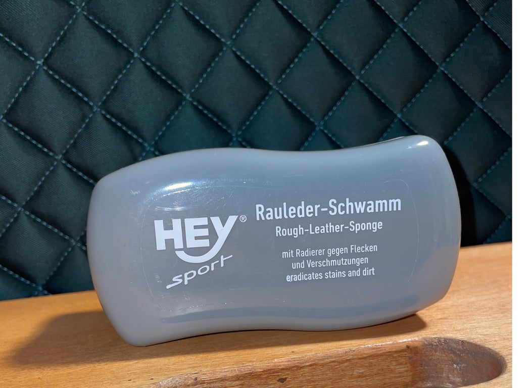 Grå rengøringssvamp til læder med teksten "HEY sport Rauleder-Schwamm Rough-Leather-Sponge" stående på et træbord foran en quiltet mørk baggrund.