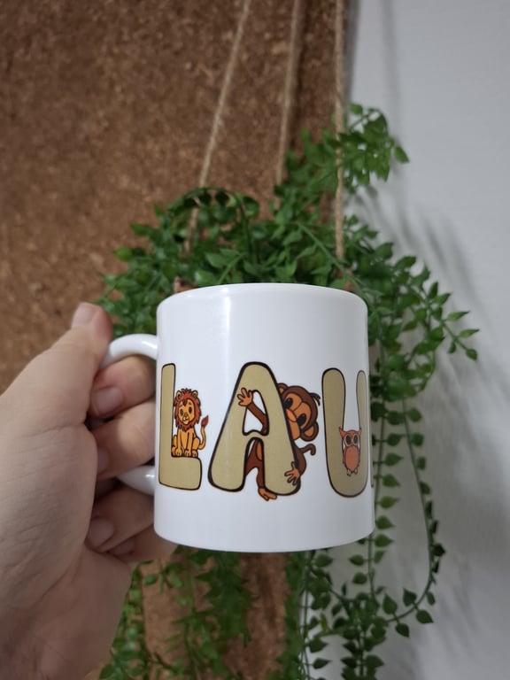 Hånd, der holder en hvid kop med ordet "LAVU" dekoreret med tegnede dyr, foran en grøn hængeplante.