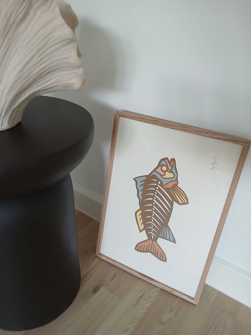 Et fotografi af en indrammet plakat med en minimalistisk, farverig fisk illustreret i brune, blå, gule og rosa nuancer, placeret på et lyst trægulv og lænet op ad en hvid væg ved siden af et sort rundt bord med en dekorativ, bølget, hvid skulptur. (2)