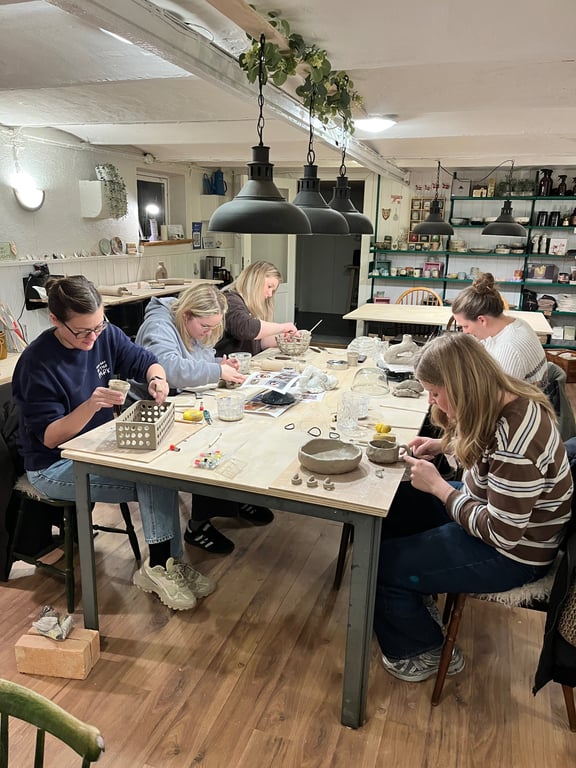 Fem kvinder sidder rundt om et bord i et lyst værksted og arbejder koncentreret med ler og keramik.