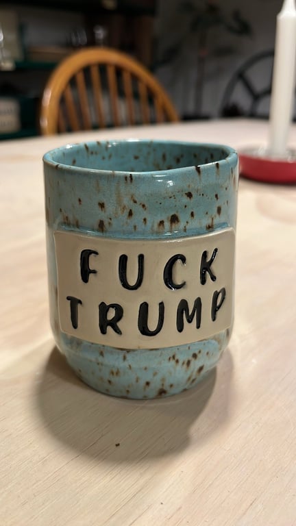 Blå keramik kop med teksten "FUCK TRUMP" i sorte bogstaver på beige firkant, placeret på et lyst bord med en træstol og stearinlys i baggrunden.
