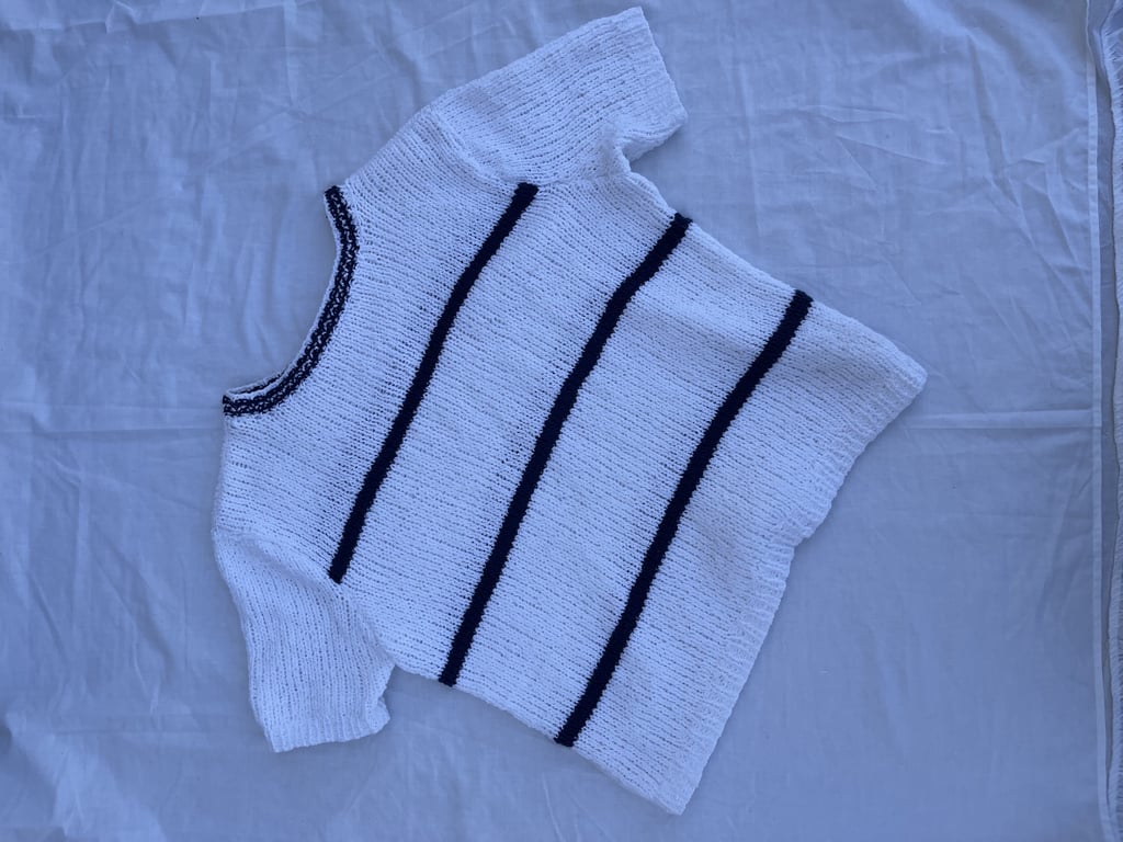 Hvid strikket kortærmet trøje med tre sorte lodrette striber og sort kant ved halsen lagt på hvidt stof.