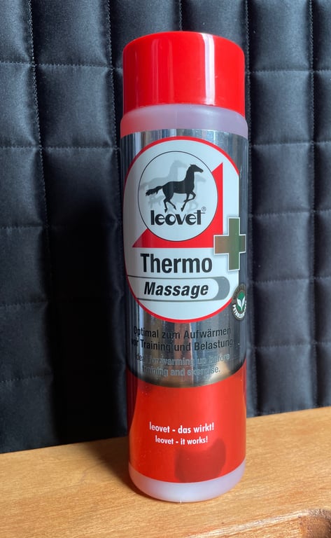 Flaske med Leovet Thermo Massage, et varmefremmende massageprodukt til træning og belastning, på en træbordplade foran en sort polstret baggrund.
