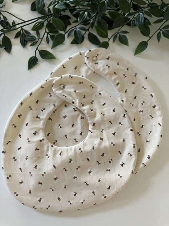 To baby savlesmække i cremefarvet stof med lille mønster af sorte og brune myrer, lagt på hvidt bord med grønne blade i baggrunden.