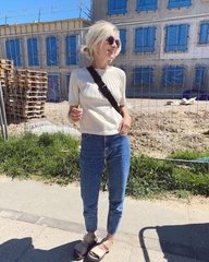 Kvinde iført solbriller, lyserød t-shirt, blå jeans og sorte sandaler, står udenfor foran en byggeplads med stabler af paller og en bygning i baggrunden. (1)