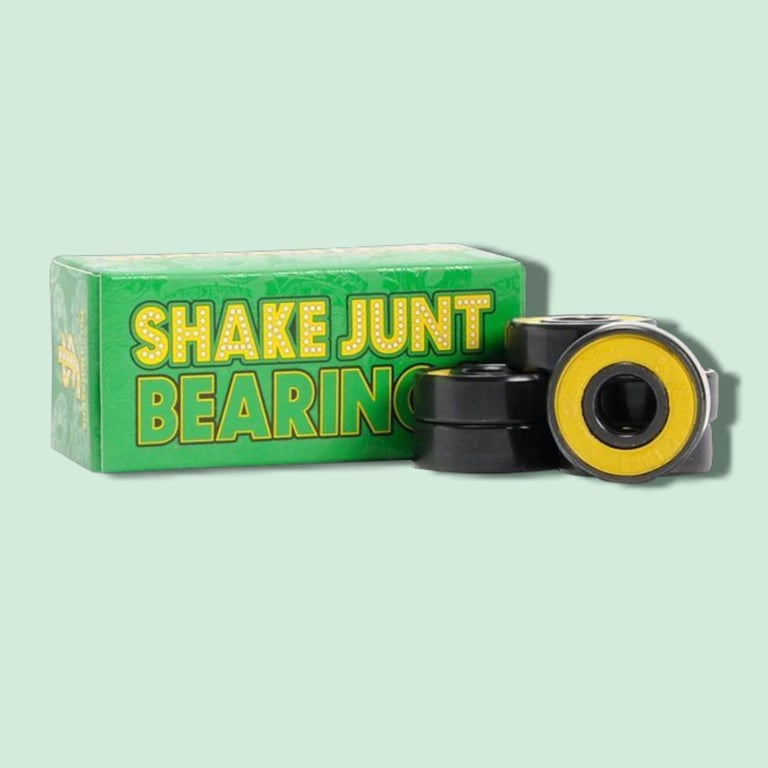 Grøn boks med teksten "Shake Junt Bearings" og fire sorte kuglelejer med gul detalje, arrangeret ved siden af boksen.