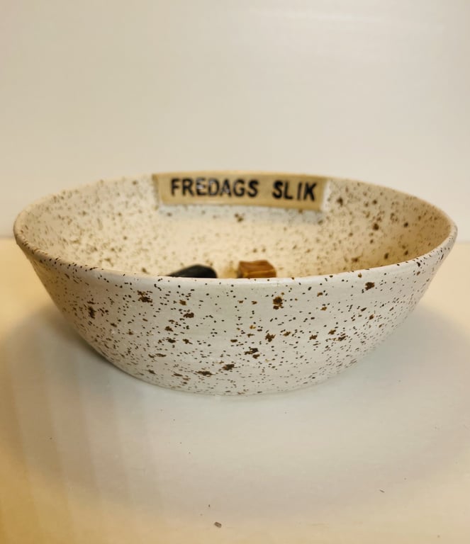 Skål med brune pletter og teksten "FREDAGS SLIK" fyldt med slik på hvid overflade.