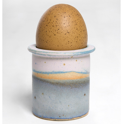 En brun æg, der står i en dekorativ, glaseret kop med blå, orange og gule nuancer på en hvid baggrund.