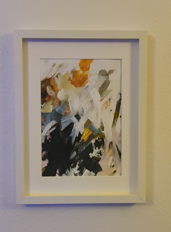 Abstrakt maleri med blandede farver, herunder hvid, orange, sort, blå og grøn, indrammet i en hvid ramme og hængt på en lys væg.