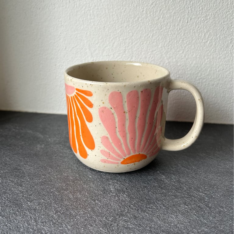 Keramikkrus med store blomster i orange og rosa på hvid baggrund, placeret på mørk bordplade foran hvid væg.