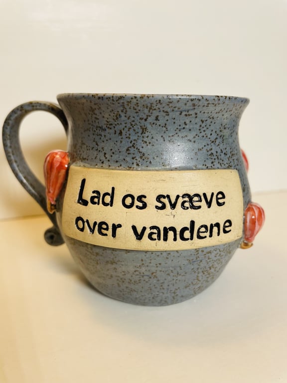 Grå keramikkop med sort skrift på beige plade, hvor der står "Lad os svæve over vandene", med små røde hjerter på siderne.