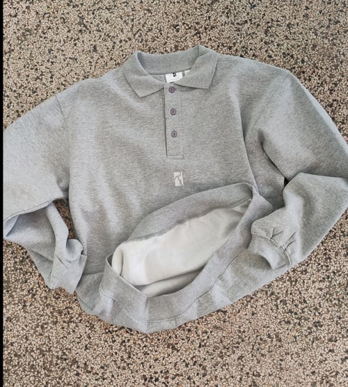 Grå sweatshirt med krave og knapper, lagt ud fladt på en pebble- eller stenmønstret overflade. (2)