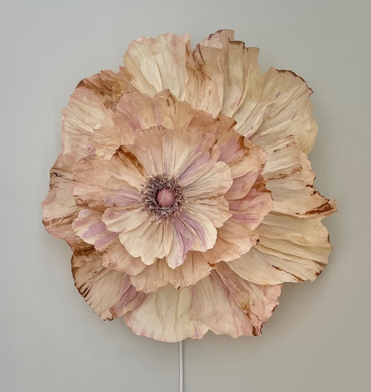 Stor, dekorativ blomst lavet af lyse papirblade med nuancer af beige, rosa og brune kanter, monteret på en væg.