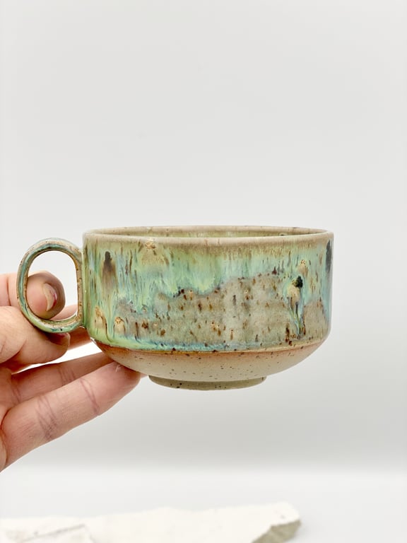 Hånd der holder en keramisk kop med grøn, blå og beige glasur mod en hvid baggrund.