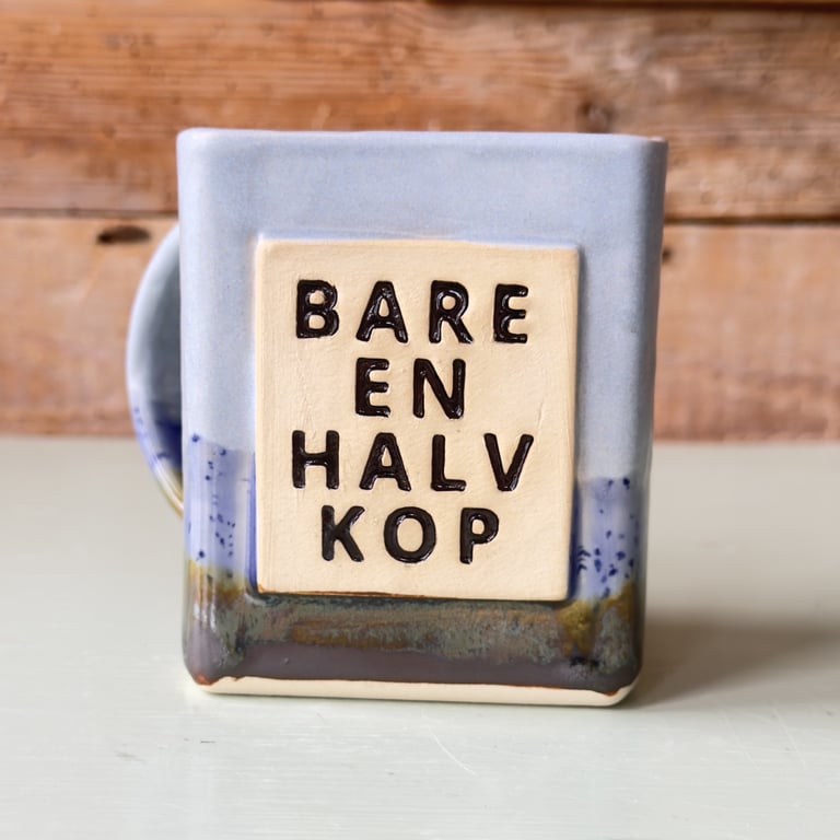 En keramisk kop med teksten "BARE EN HALV KOP" i sort relief på en lys firkant, koppen har blå og brun glasur, står på et lyst bord med træbaggrund.