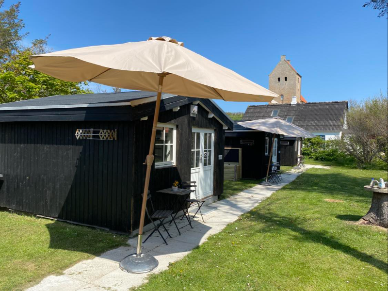 To huse i sort træ med hvide døre og vinduer, to beige parasoller, små borde og stole på en græsplæne med en kirke i baggrunden under en klar blå himmel.