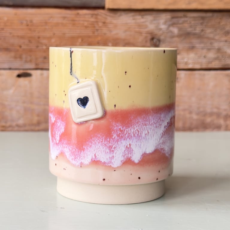 Keramikkrus med gul, pink og hvid glasur og en lille tebrevs-figur med et sort hjerte på side.