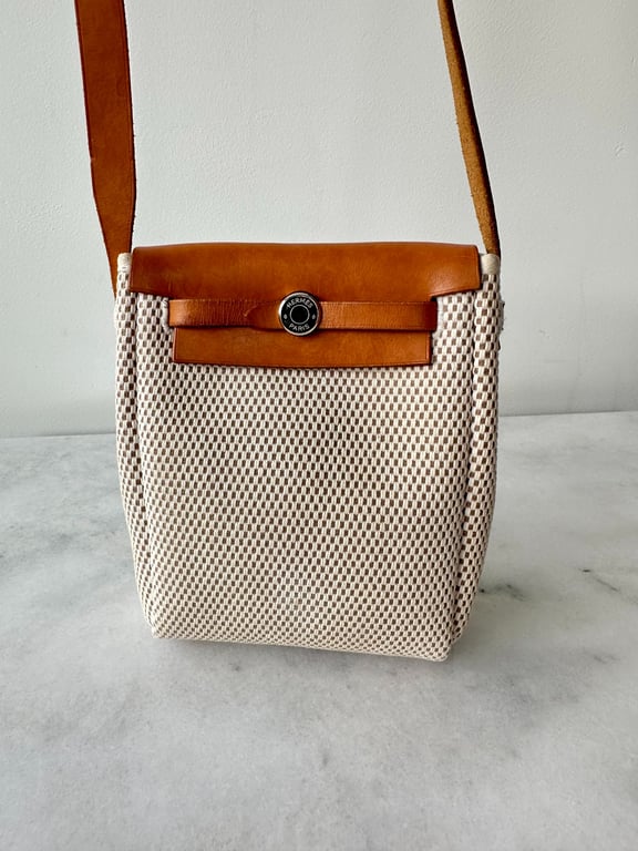 Beige og lysebrun Hermes skuldertaske med flettet mønster og læderdetaljer, placeret på en marmoroverflade.