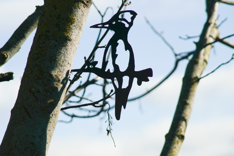 Silhuet af en fuglefigur i metal ophængt på en gren mod en blå himmel.