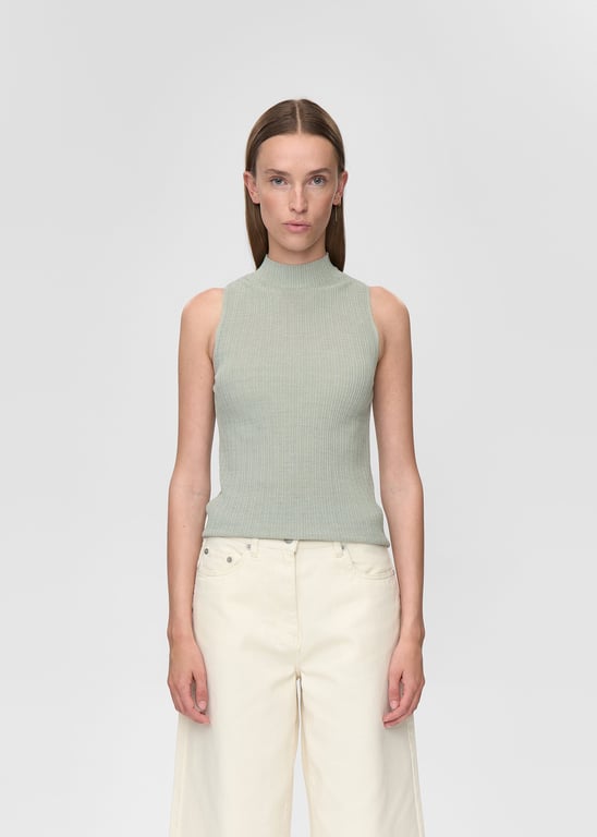 Kvinde iført lysegrøn ribstrikket ærmeløs top og cremefarvede bukser, stående mod en lys baggrund.