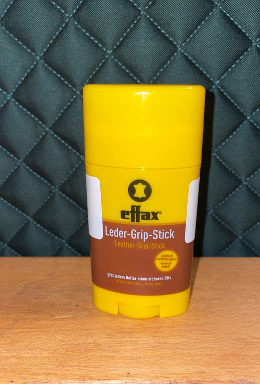 Gul og brun plastbeholder med tekst "effax Leder-Grip-Stick" stående på træbord med mørkt quiltet stof baggrund.