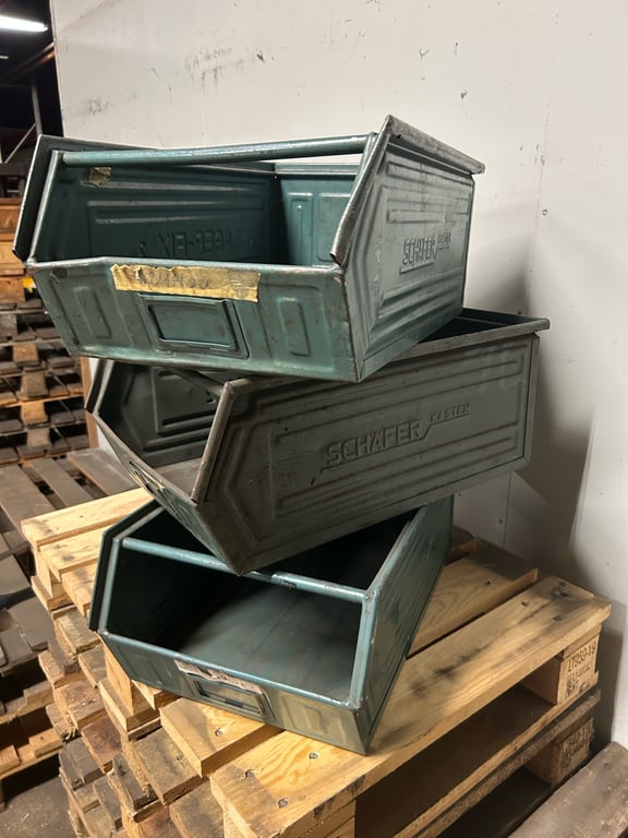 Tre blåmetal opbevaringskasser stablet oven på hinanden på en træpalle i et lagerområde.