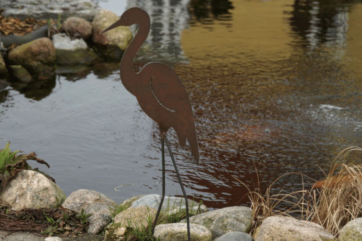 Rustfarvet metalfigur af en fugl ved kanten af en dam med sten og græs.
