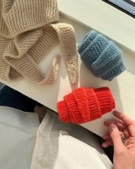 Tre strikkede grydelapper i beige, blå og rød ligger på en hvid overflade, mens en hånd rører ved den røde. (1)