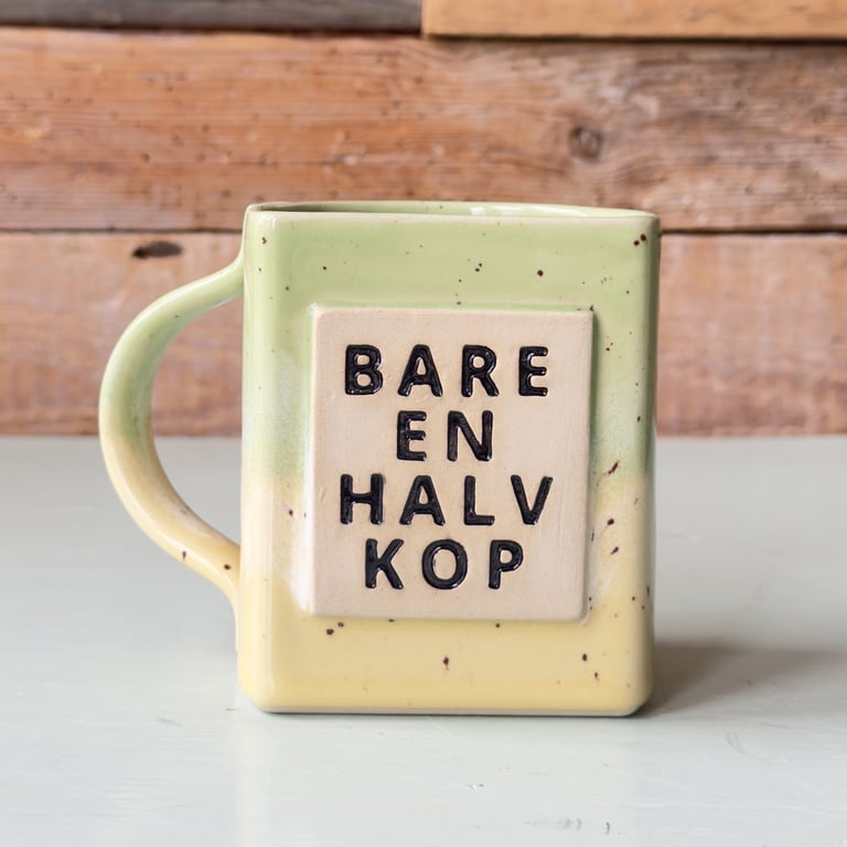 En firkantet, gul og grøn ceramikkop med teksten "BARE EN HALV KOP" i sorte bogstaver på forsiden, placeret på en hvid overflade med en trævæg i baggrunden.