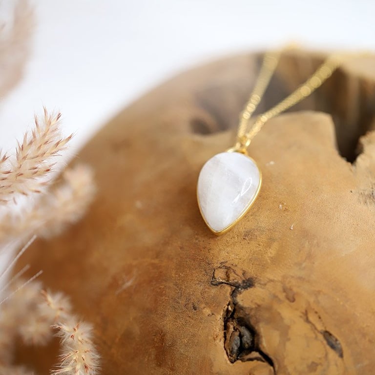 Nærbillede af en guldfarvet halskæde med vedhæng i hvid sten, lagt på en rund trægenstand med tørrede blomster i baggrunden. (3)