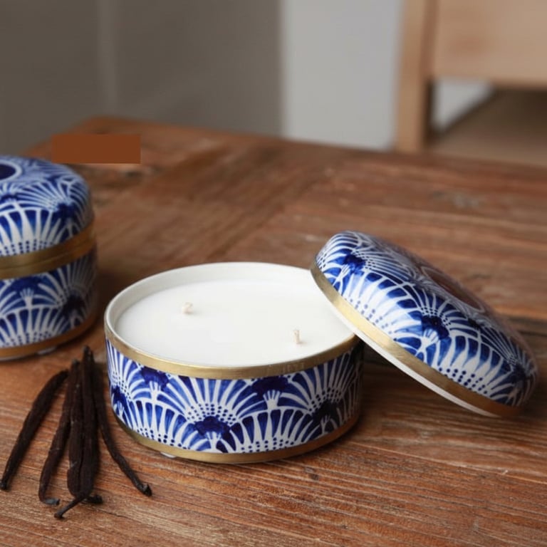 To lys i en rund, blå og hvid mønstret keramisk beholder med låg, placeret på et træbord ved siden af hele vaniljestænger.
