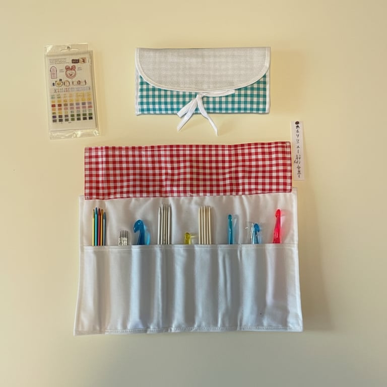 Sæt med hæklenåle og strikkepinde i et hvidt etui med rød ternet foring, sammenfoldet etui med blå tern og en pakke med broderitråd.