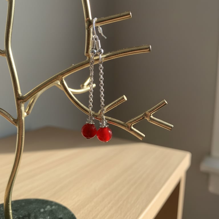 Sølvøreringe med kæde og røde perler hængende på en guldgrentallerken på et lyst træbord.