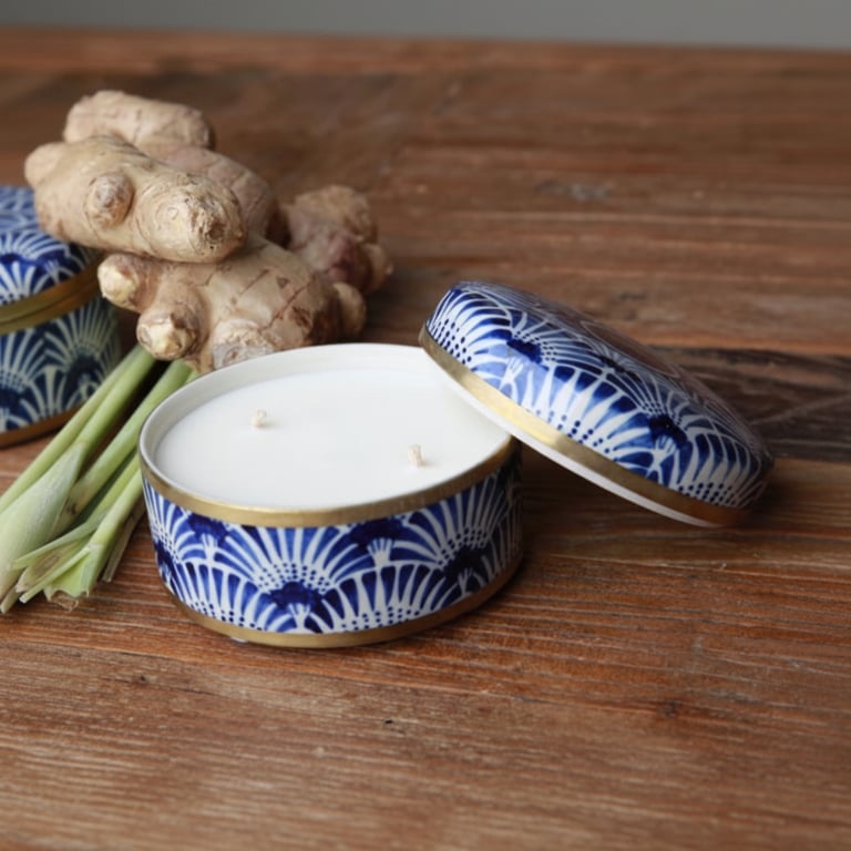 Lys i en blå og hvid mønstret tinbeholder med låg, placeret på et træbord ved siden af frisk ingefær og citrongræs.